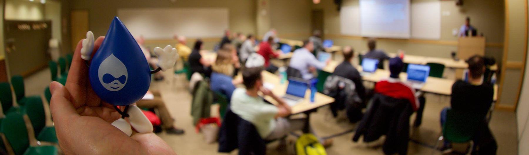 In the foreground, a hand holding a blue foam Druplicon figurine. In the background, attendees watch a session in progress.